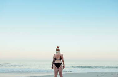 Unerkennbare weibliche Winterbadende am Strand in Badekleidung. Rückansicht einer anonymen Frau, die im Winter auf das Meerwasser schaut. Winterbadende, die allein am Meer steht. - JLPSF08870