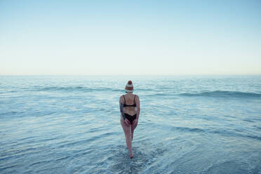 Anonyme Frau, die am Strand ins kalte Meerwasser steigt. Rückansicht einer nicht erkennbaren Winterbadenden in Badekleidung und mit Wollmütze. Frau, die ein Winterbad am Meer nimmt. - JLPSF08884