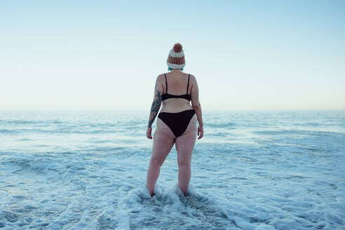 Female winter bather watching the sea waves at the beach. Rearview of an unrecognisable winter bather wearing swimwear and a wooly hat. Woman standing in the cold sea water. - JLPSF08887