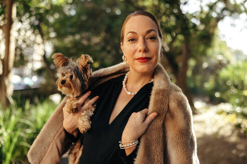 Elegant woman looking at the camera while holding her puppy outdoors. High class woman standing alone in a park during the day. Female pet owner going for a walk with her dog. - JLPSF08894