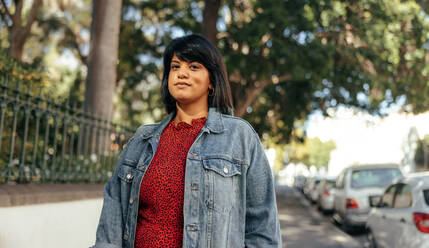 Woman looking at the camera while standing on a sidewalk in the city. Mid-adult woman standing alone next to a park outdoors. Confident mid-adult woman wearing a denim jacket. - JLPSF08915