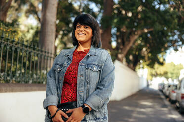 Happy mid-adult woman smiling at the camera while standing on a sidewalk in the city. Cheerful woman standing alone next to a park outdoors. Confident mid-adult woman wearing a denim jacket. - JLPSF08916