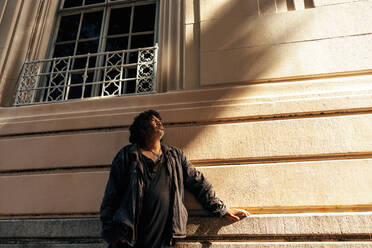 Homeless man looking up thoughtfully while standing alone outdoors during the day. Middle aged underprivileged man having a reflective moment while standing against a brick wall in the city. - JLPSF08926