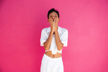 Happy queer teenager coming out and embracing his identity. Cheerful gay teenager covering his mouth with his hands while smiling. Young teenage boy standing against a pink background. - JLPSF08955