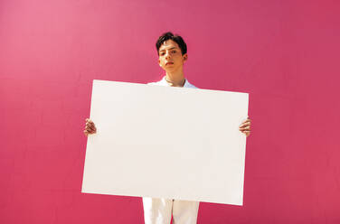 Selbstbewusster queerer Junge, der ein leeres Plakat vor einem rosafarbenen Hintergrund hält. Selbstbewusster jugendlicher Aktivist, der ein weißes Transparent in einem Studio zeigt. Junger schwuler Junge, der sich outet und seine Identität annimmt. - JLPSF08958