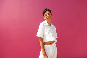 Happy teenage boy standing against a pink background. Cheerful young queer boy smiling at the camera while standing in a studio. Confident gay teenager coming out and embracing his identity. - JLPSF08966