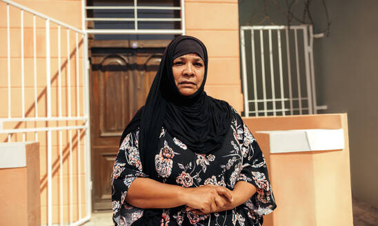 Selbstbewusste muslimische Frau, die in die Kamera schaut, während sie tagsüber vor ihrem Haus steht. Selbstbewusste Frau mittleren Alters, die einen Hijab trägt und allein steht. - JLPSF08969