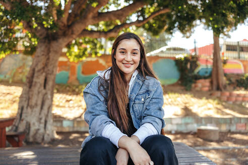 Unbekümmertes Teenager-Mädchen, das fröhlich in die Kamera lächelt. Glückliches brünettes Mädchen in Jeansjacke im Freien in der Stadt. Weibliche Jugendliche, die tagsüber allein in einem Stadtpark sitzt. - JLPSF08976