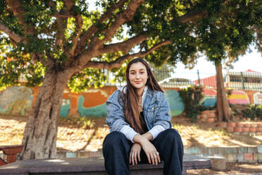 Self-confident teenage girl looking at the camera outdoors. Carefree brunette girl wearing a denim jacket in the city. Female youngster sitting alone in an urban park during the day. - JLPSF08979