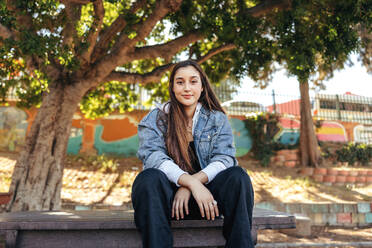 Young teenage girl looking at the camera outdoors. Confident brunette girl wearing a denim jacket in the city. Female youngster sitting alone in an urban park during the day. - JLPSF08980