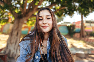 Adorable teenage girl looking at the camera outdoors. Carefree young brunette girl wearing a denim jacket in the city. Female youngster sitting alone in an urban park during the day. - JLPSF08984