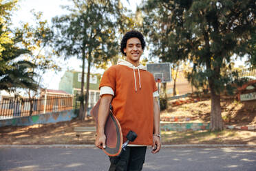 Cheerful skateboarder holding his skateboard in an urban park. Happy teenage boy smiling at the camera while standing outdoors. Sporty teenager wearing casual clothing during the day. - JLPSF08993