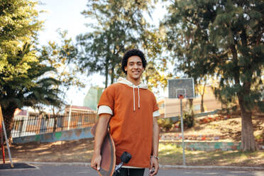 Happy skateboarder carrying his skateboard outdoors during the day. Cheerful teenage boy smiling at the camera while standing outdoors. Sporty teenager wearing casual clothing during the day. - JLPSF08994