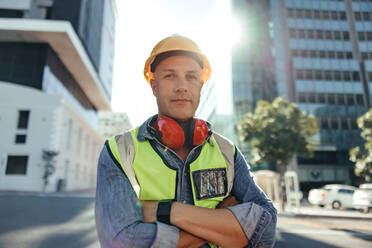 Selbstbewusster Bauarbeiter, der im Freien in die Kamera schaut. Mittelgroßer Bauingenieur, der mit verschränkten Armen in der Stadt steht. Arbeiter, der vor einem Hochhaus steht. - JLPSF09000