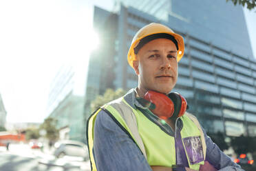 Confident workman looking at the camera while standing with his arms crossed in the city. Mid-adult construction worker standing in front of high rise buildings in his workwear. - JLPSF09001
