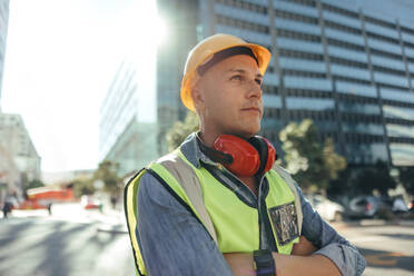 Construction worker looking away thoughtfully. Mid-adult workman standing with his arms crossed in the city. Blue collar worker standing in front of high rise buildings in his work clothes. - JLPSF09002