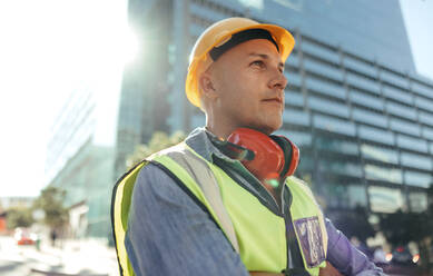 Arbeiter, der mit verschränkten Armen in der Stadt steht und nachdenklich in die Ferne blickt. Bauarbeiter im mittleren Erwachsenenalter, der in seiner Arbeitskleidung vor einem Hochhaus steht. - JLPSF09003