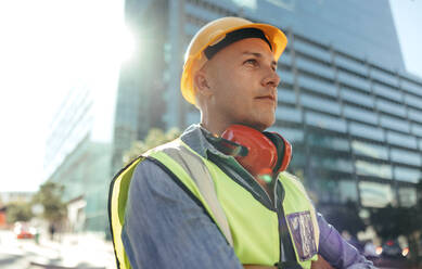 Blue collar worker looking away thoughtfully while standing with his arms crossed in the city. Mid-adult construction worker standing in front of high rise buildings in his workwear. - JLPSF09003