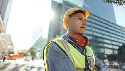 Nachdenklicher Arbeiter, der in der Stadt steht. Mittelgroßer Bauarbeiter, der nachdenklich wegschaut, während er mit verschränkten Armen im Freien steht. Arbeiter, der vor einem Hochhaus steht. - JLPSF09004