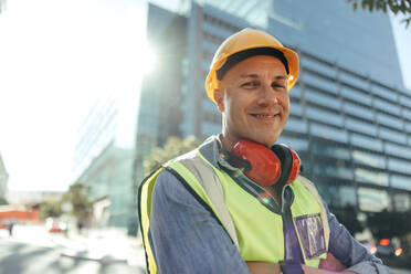 Glücklicher Bauarbeiter, der in die Kamera lächelt, während er mit verschränkten Armen in der Stadt steht. Mittelgroßer Arbeiter, der in seiner Arbeitskleidung vor einem Hochhaus steht. - JLPSF09005