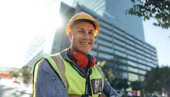Fröhlicher Bauarbeiter, der in die Kamera lächelt, während er mit verschränkten Armen in der Stadt steht. Mittelgroßer Arbeiter, der in seiner Arbeitskleidung vor einem Hochhaus steht. - JLPSF09006