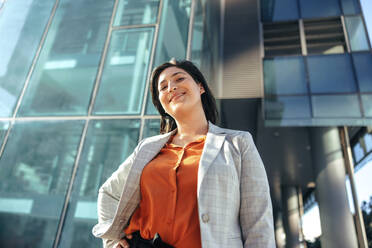 Fröhliche Geschäftsfrau, die in die Kamera lächelt, während sie vor einem Bürohochhaus in der Stadt steht. Erfolgreiche Unternehmerin, die allein vor ihrem Arbeitsplatz steht. - JLPSF09009