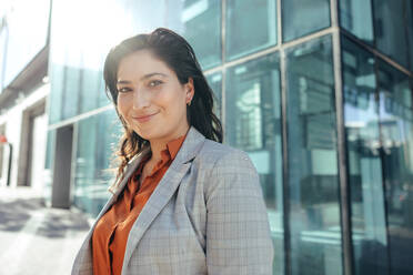 Happy businesswoman smiling at the camera while standing in front of a high rise office building in the city. Successful female entrepreneur standing alone outside her workplace. - JLPSF09013