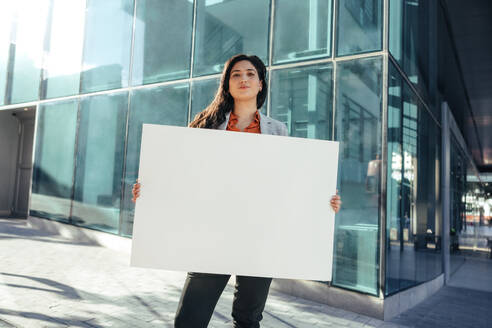 Confident businesswoman holding a blank banner and looking at the camera while standing in front of a high rise office building in the city. Business activist standing alone outside her workplace. - JLPSF09019