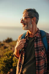 Glücklicher reifer Wanderer, der fröhlich wegschaut, während er mit seinem Rucksack auf einem Hügel steht. Abenteuerlustiger männlicher Rucksacktourist, der eine Wanderung bei Sonnenuntergang genießt. - JLPSF09032
