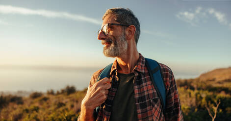 Enjoying the hilltop views. Happy mature hiker looking away with a smile on his face while standing on top of a hill with a backpack. Adventurous backpacker enjoying a hike at sunset. - JLPSF09033