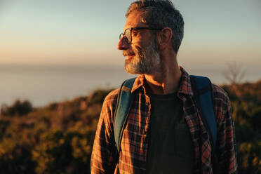 Adventurous male hiker looking away while standing on a hilltop. Happy mature hiker standing with a backpack on outdoors. Cheerful mature man enjoying the sunset and view. - JLPSF09036