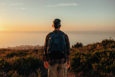 Unbekannter Rucksacktourist mit Panoramablick auf einer Bergkuppe. Rückansicht eines männlichen Wanderers, der allein auf einem Küstenhügel steht. Abenteuerlustiger reifer Mann, der den Sonnenuntergang im Freien genießt. - JLPSF09040