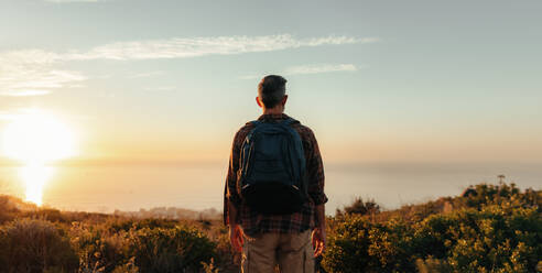 Rearview of a backpacker looking at the panoramic view on a hilltop. Unrecognisable male hiker standing alone on a coastal hill. Adventurous mature man enjoying the sunset outdoors. - JLPSF09041