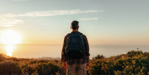 Rückansicht eines Rucksacktouristen, der auf einer Bergkuppe den Panoramablick genießt. Unbekannter männlicher Wanderer, der allein auf einem Küstenhügel steht. Abenteuerlustiger reifer Mann, der den Sonnenuntergang im Freien genießt. - JLPSF09041