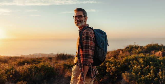 Glücklicher männlicher Wanderer, der in die Kamera lächelt, während er alleine steht. Fröhlicher reifer Mann, der einen Rucksack trägt und auf einer Bergkuppe steht. Abenteuerlustiger Rucksacktourist, der eine Wanderung bei Sonnenuntergang genießt. - JLPSF09042