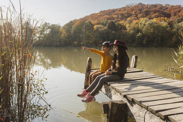 Boy pointing to girl sitting together on jetty at lake - OSF01069