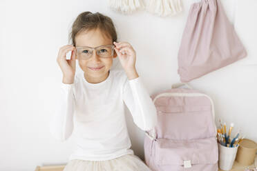 Lächelndes Mädchen mit Brille sitzt mit Rucksack auf dem Tisch - SSYF00010