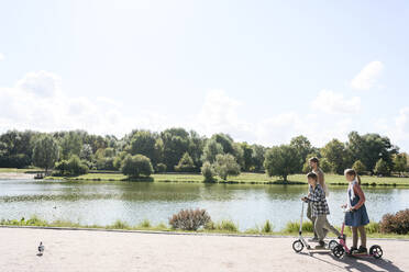 Brother and sister riding push scooter by father walking in park on weekend - EYAF02178