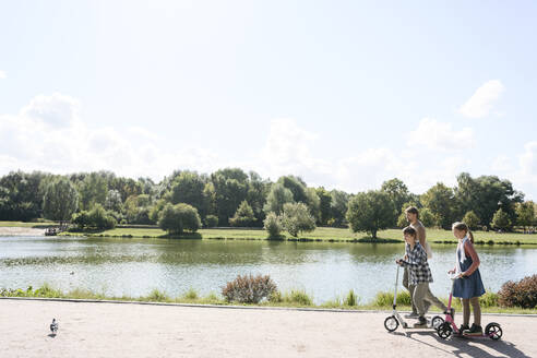 Bruder und Schwester fahren Roller, während ihr Vater am Wochenende im Park spazieren geht - EYAF02178