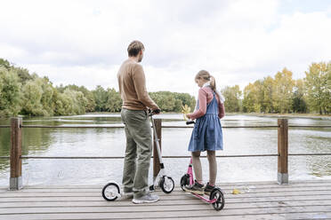 Vater und Tochter mit Tretrollern auf dem Steg über den See im Park - EYAF02181