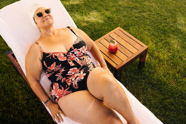 Ältere Frau entspannt sich auf einer Liege im Badeanzug. Unbeschwerte ältere Frau, die ihren Sommerurlaub in einem Luxus-Spa-Resort genießt. Glückliche reife Frau, die sich nach der Pensionierung amüsiert. - JLPSF09118