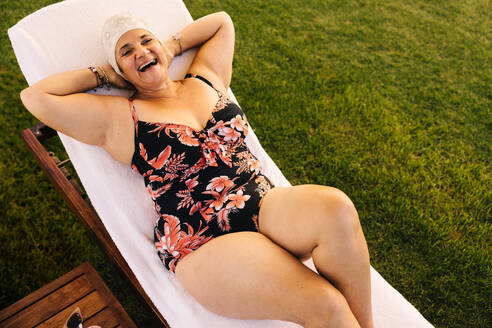 Fröhliche ältere Frau, die sich im Badeanzug auf einer Liege entspannt. Unbeschwerte ältere Frau, die im Urlaub in einem Luxus-Spa-Resort fröhlich lacht. Reife Frau, die sich nach der Pensionierung amüsiert. - JLPSF09120