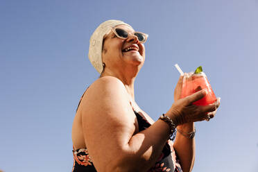 Fröhliche ältere Frau, die einen Tiki-Cocktail in Badekleidung trinkt. Unbeschwerte ältere Frau, die während eines Sommerurlaubs fröhlich lächelt. Glückliche reife Frau, die sich nach der Pensionierung amüsiert. - JLPSF09127