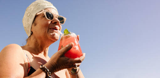 Elderly woman drinking a tiki cocktail in swimwear. Carefree senior woman enjoying her summer vacation at a tropical spa resort. Happy mature woman enjoying herself after retirement. - JLPSF09128