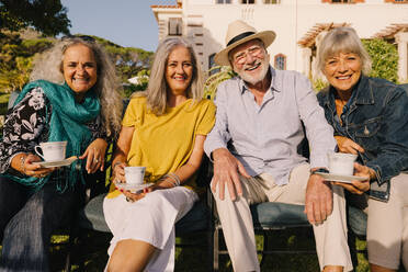 Happy senior citizens smiling at the camera while relaxing outside a retirement home. Group of carefree elderly people having tea together and enjoying their golden years. - JLPSF09149