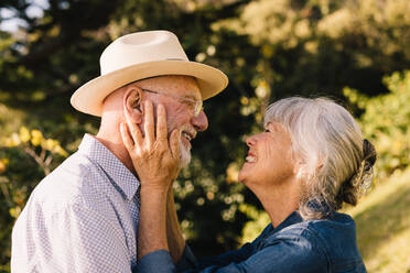 Cheerful senior couple smiling at each other with love and affection. Grey-haired couple sharing a romantic moment while standing in a park. Happy elderly couple adoring each other outdoors. - JLPSF09153