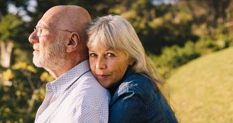 Beautiful elderly couple embracing each other in a park. Romantic senior woman looking at the camera while standing behind her husband. Mature couple spending quality time after retirement. - JLPSF09158