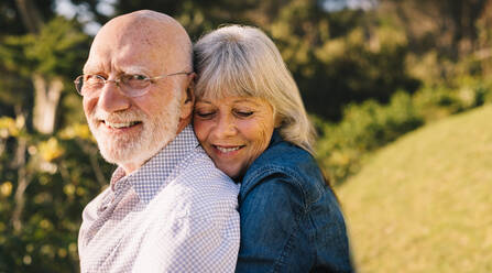 Glückliches reifes Paar, das sich umarmt, während es in einem Park steht. Romantisches älteres Paar, das lächelt und zusammen die Sonne genießt. Liebevolles älteres Paar, das nach dem Ruhestand Zeit miteinander verbringt. - JLPSF09159