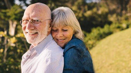 Happy mature couple embracing each other while standing in a park. Romantic elderly couple smiling and enjoying the sun together. Affectionate senior couple spending quality time after retirement. - JLPSF09159