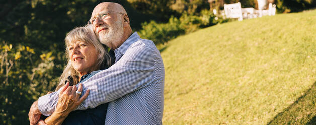 Elderly couple embracing each other while standing in a park. Romantic senior couple enjoying the sun in each other's arms. Mature couple spending quality time after retirement. - JLPSF09162